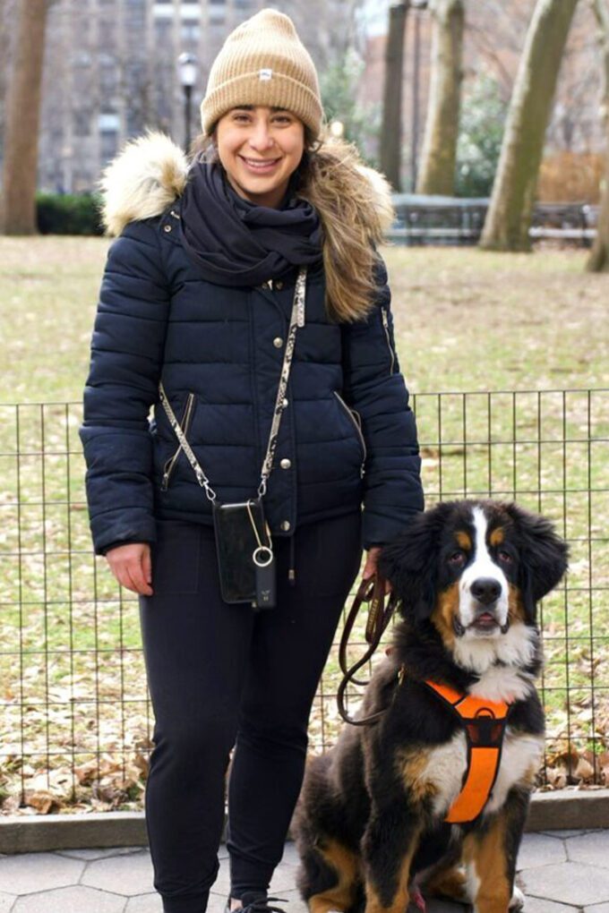 A woman in a tan beanie, navy winter coat, and black pants smiles while standing next to a large black, brown, and white dog wearing an orange harness in a park with bare trees, showcasing positive training and behavior skills.