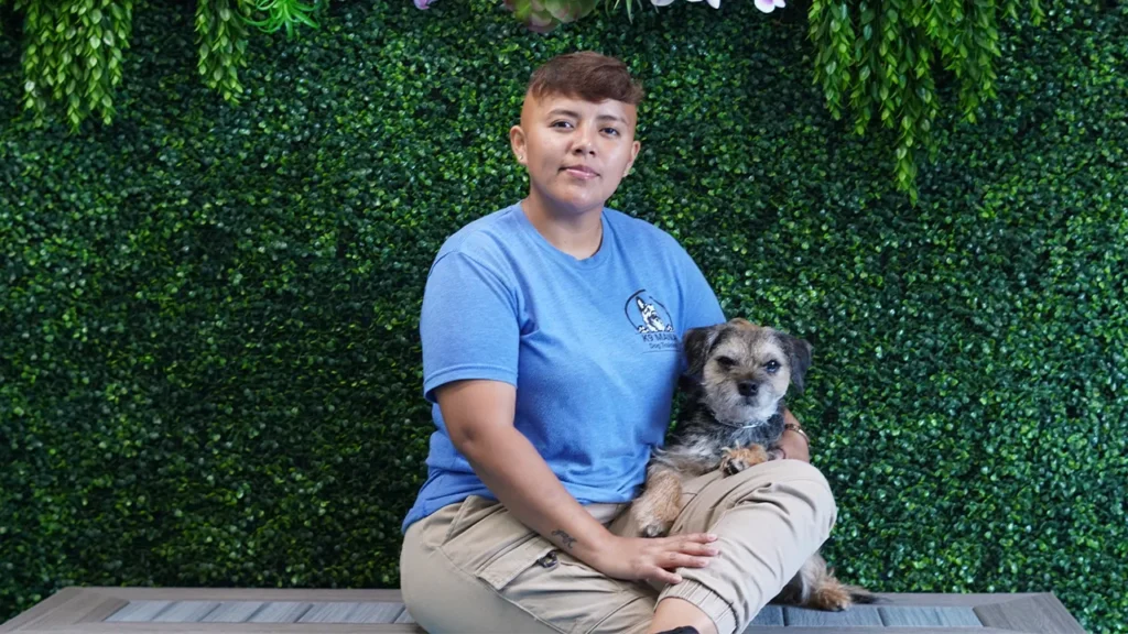 A person in a blue shirt and khaki pants sits on a bench, holding a small, scruffy dog known for its good obedience. They are in front of a backdrop covered with green foliage and hanging plants.