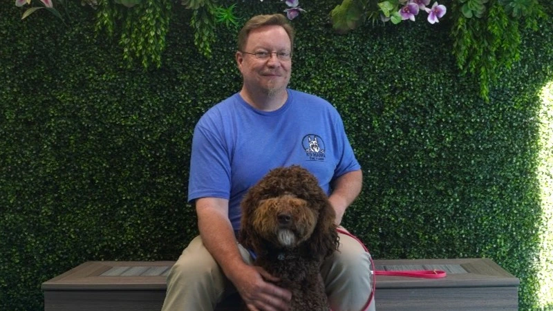 A man in a blue T-shirt sits on a bench in front of a green leafy wall, smiling at the camera, with his curly-haired brown puppy sitting obediently in front of him on a pink leash.