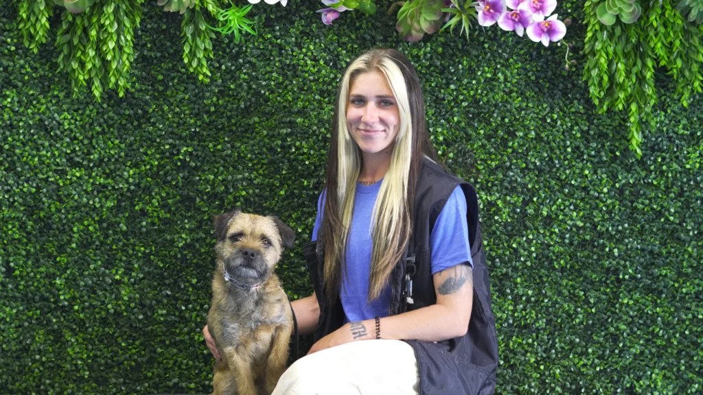 A woman with long blonde and brown hair sits beside her small brown dog, both facing the camera, in front of a lush green wall decorated with hanging plants and purple orchids—a perfect moment of companionship and obedience.