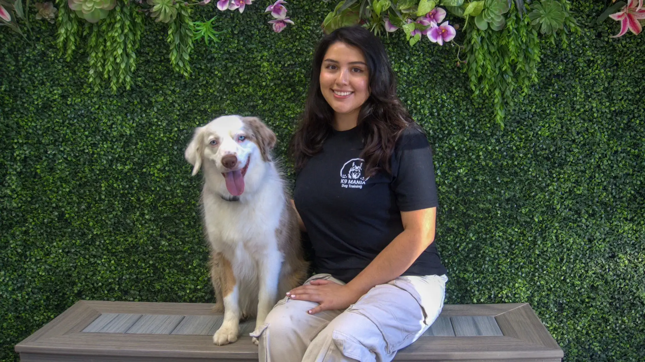A woman sits on a bench next to a happy white and brown canine with its tongue out. They are in front of a green leafy wall decorated with pink and purple flowers, enjoying a break after some training.