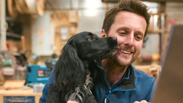 A man smiles as a playful puppy licks his face in a workshop setting, with woodworking tools and materials visible in the background.