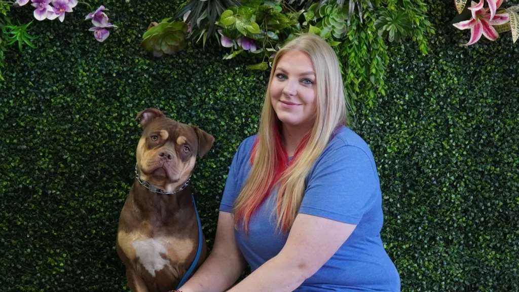 A woman with long blonde hair and a blue shirt sits next to her obedient brown canine with a white chest. They pose in front of a lush green wall decorated with flowers and plants.