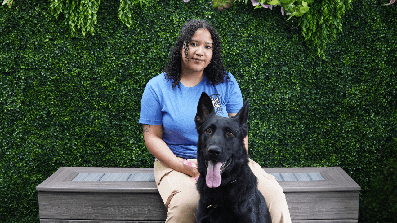 A person with curly hair wearing a blue shirt and khaki pants sits on a bench in front of a leafy green wall, while their large black dog, showing off its training and obedience, sits proudly in front, tongue out and looking at the camera.
