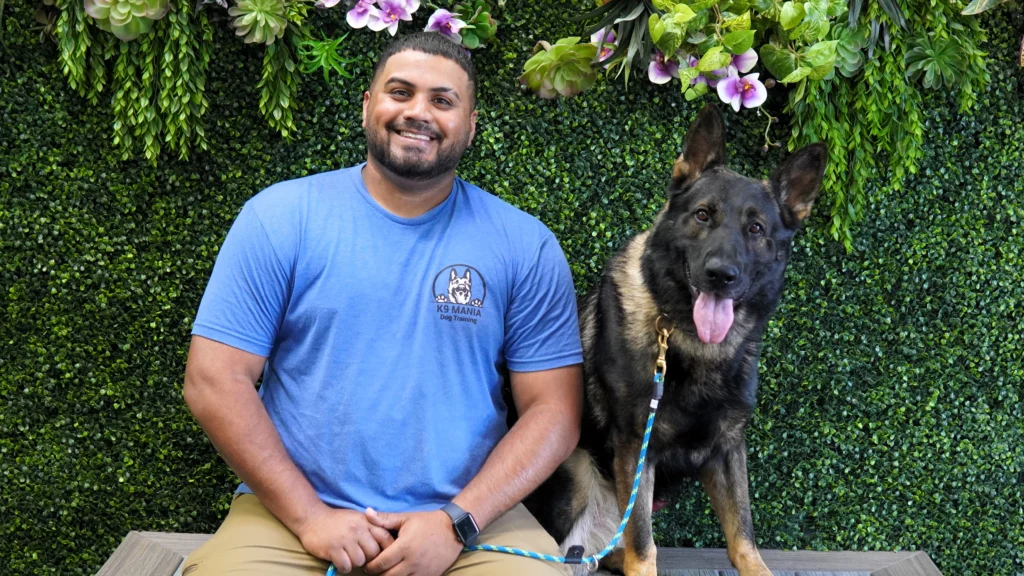 A smiling man in a blue T-shirt sits next to his large German Shepherd on a bench, the dog leashed and showing off its training. They both look at the camera, framed by a green leafy wall and purple flowers in the background.