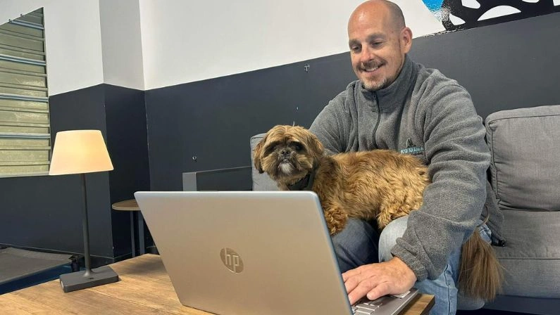 Smiling man works on laptop with a small brown dog in his lap, thinking about Long Island dog boarding.
