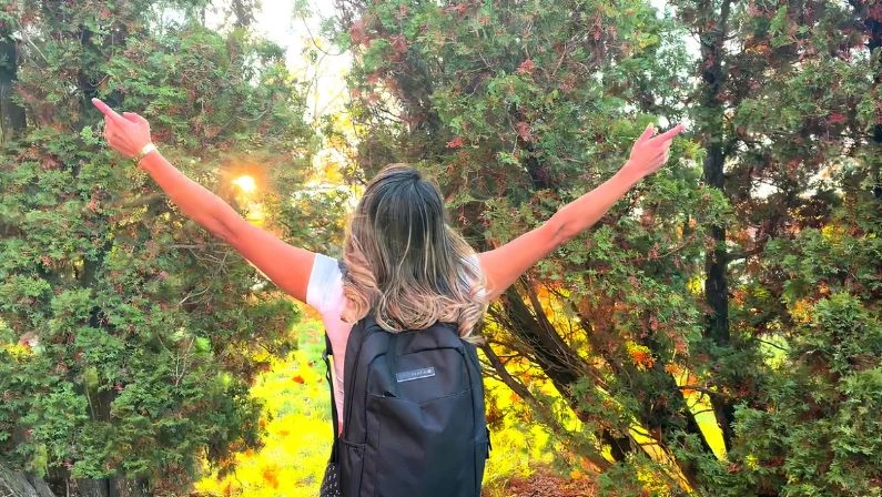 A person with a backpack stands outdoors at a dog boarding camp, facing trees with arms raised.